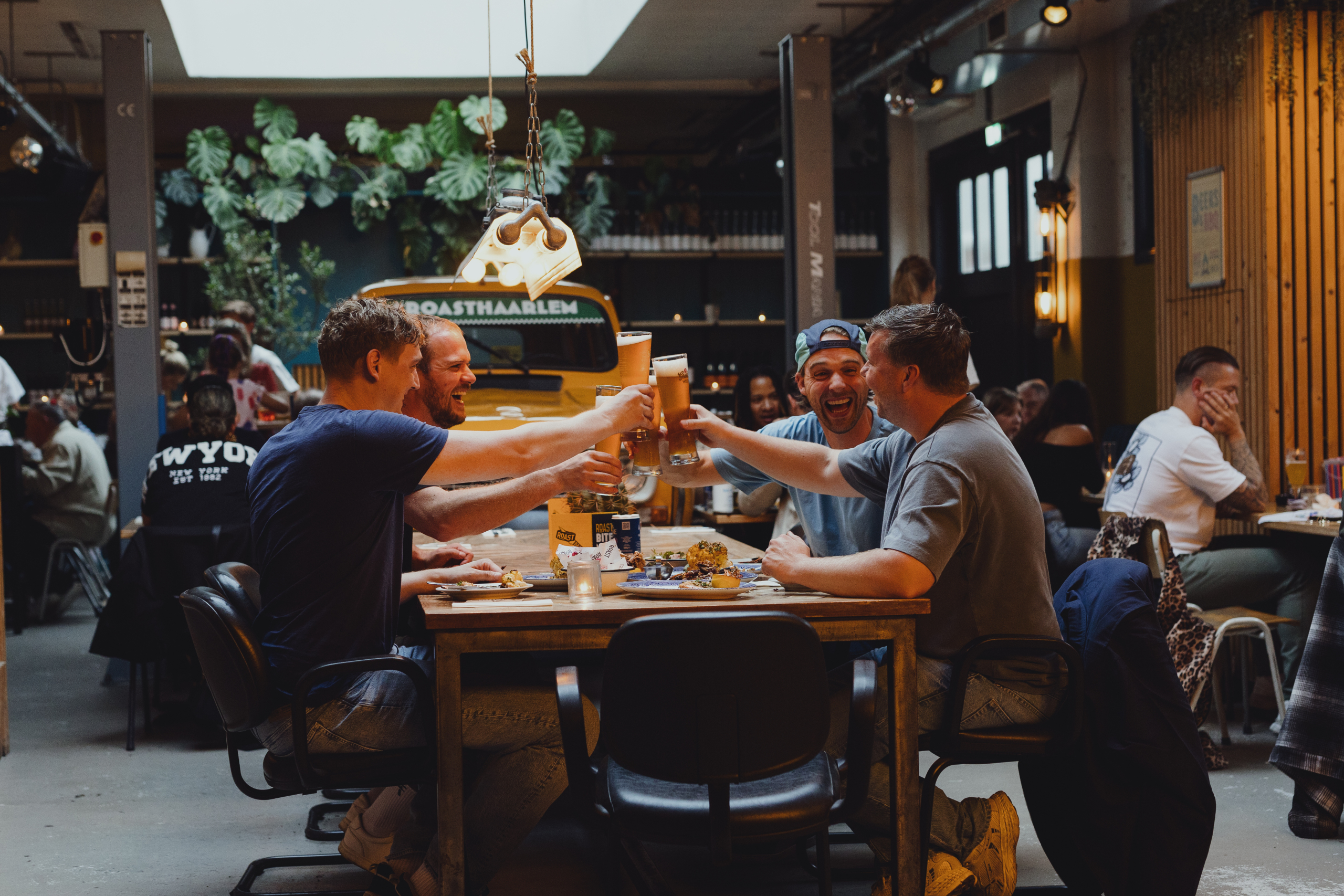 Vier mannen proosten met bier in een gezellige, industriële eetgelegenheid met groene planten en houten decor.