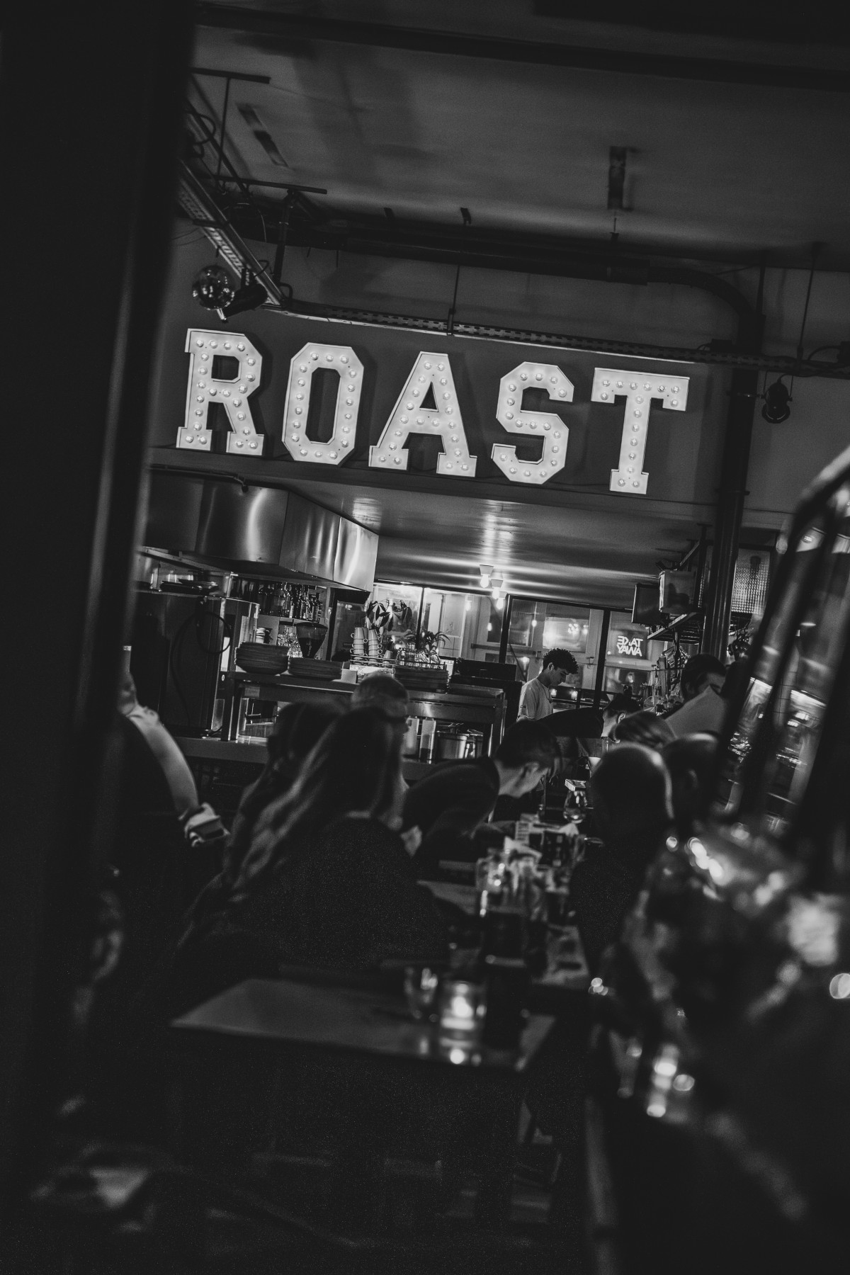 Gezellige restaurantomgeving met grote verlichte "ROAST" letters, donkere sfeer en gasten aan tafels.