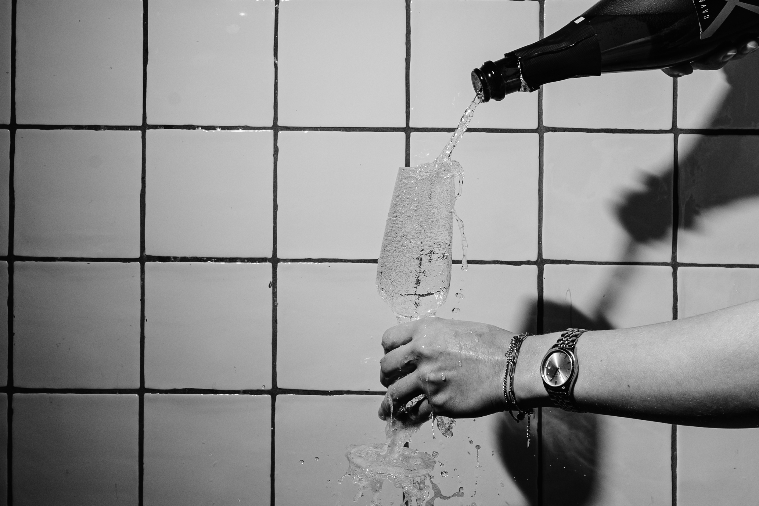 Champagne wordt in een glas geschonken tegen een achtergrond van witte tegels, hand met horloge zichtbaar.